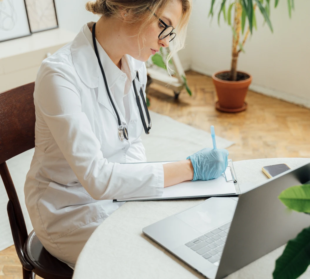 Healthcare professional writing notes while working on a laptop